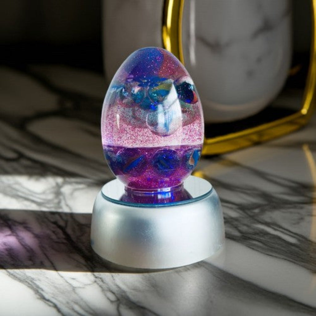 Colour Changing Egg - A decorative egg-shaped resin lamp with swirling purple, blue, and pink glitter inside, sitting on a frosted white base on a marble surface.