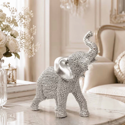 A beaded elephant figurine with a shiny silver-toned ear stands on a light marble table in a softly lit, elegant room with cream furniture and white flowers in the background.