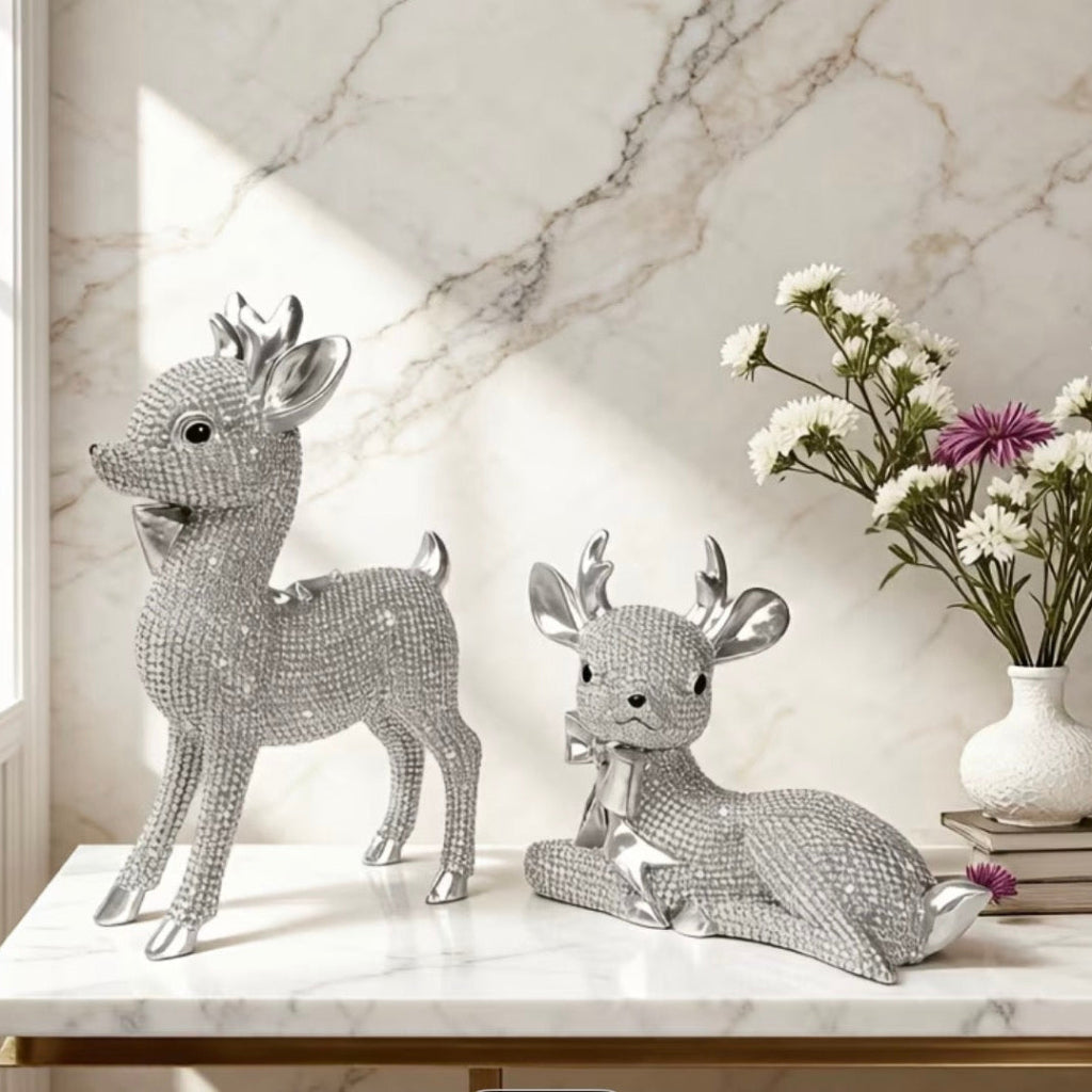 Reindeer Gift Set - Two silver deer figurines covered in sparkling rhinestones, one standing and one reclining, displayed on a white marble console table with a vase of flowers and books in the background.