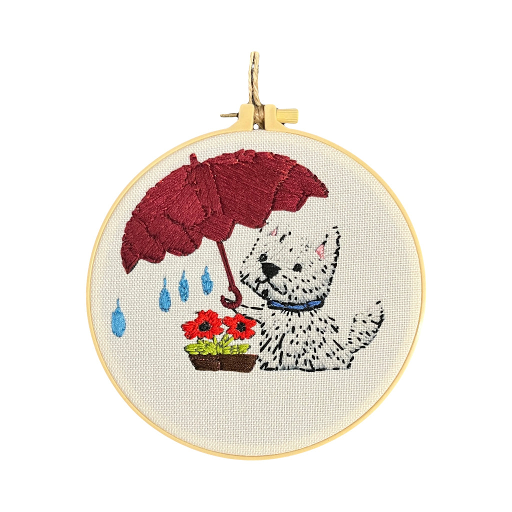 Dog Wall Art - A circular embroidery hoop containing a finished embroidered design of a white West Highland terrier dog holding a dark red umbrella under blue raindrops, with red flowers in a pot, on a white background.