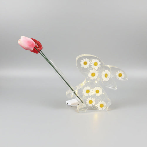 Bunny Bud Vase - A clear resin bunny figurine filled with small white daisy flowers and LED lights, with yellow centres, holding a pink tulip stem, photographed on a smooth light grey studio background.
