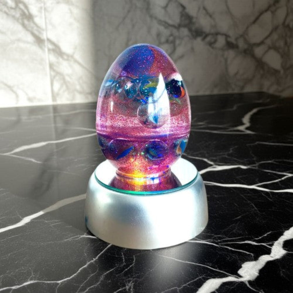 Colour Changing Egg - A colourful, glittery egg-shaped decorative object with swirling purple, pink, blue, and gold tones, displayed on a metallic silver base against a black marble-patterned surface and background.