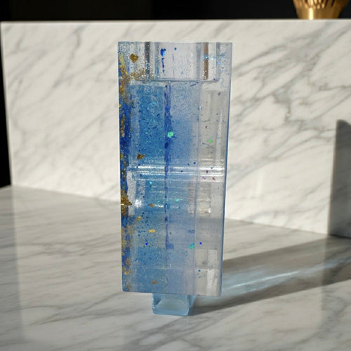 Side view of a  translucent rectangular candle holder with layered blue and clear sections, standing on a small base against a white marble-patterned background.