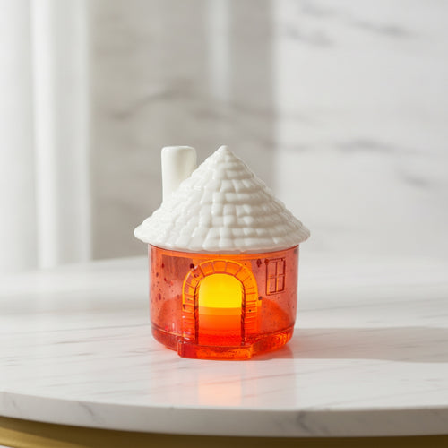 A small house-shaped candle holder with a textured white ceramic roof and a translucent orange glass body glowing with warm light, set on a white marble surface.