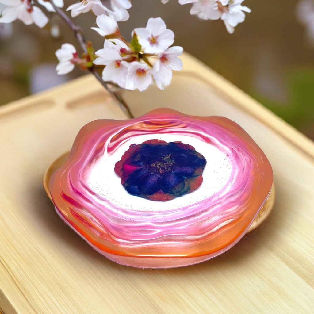 Decorative Trays - A decorative tray dish with a flower motif, featuring a glossy pink and orange outer ring, a white centre, and a dark purple flower in the middle. It is placed on a light wooden surface with blurred white blossoms in the background.