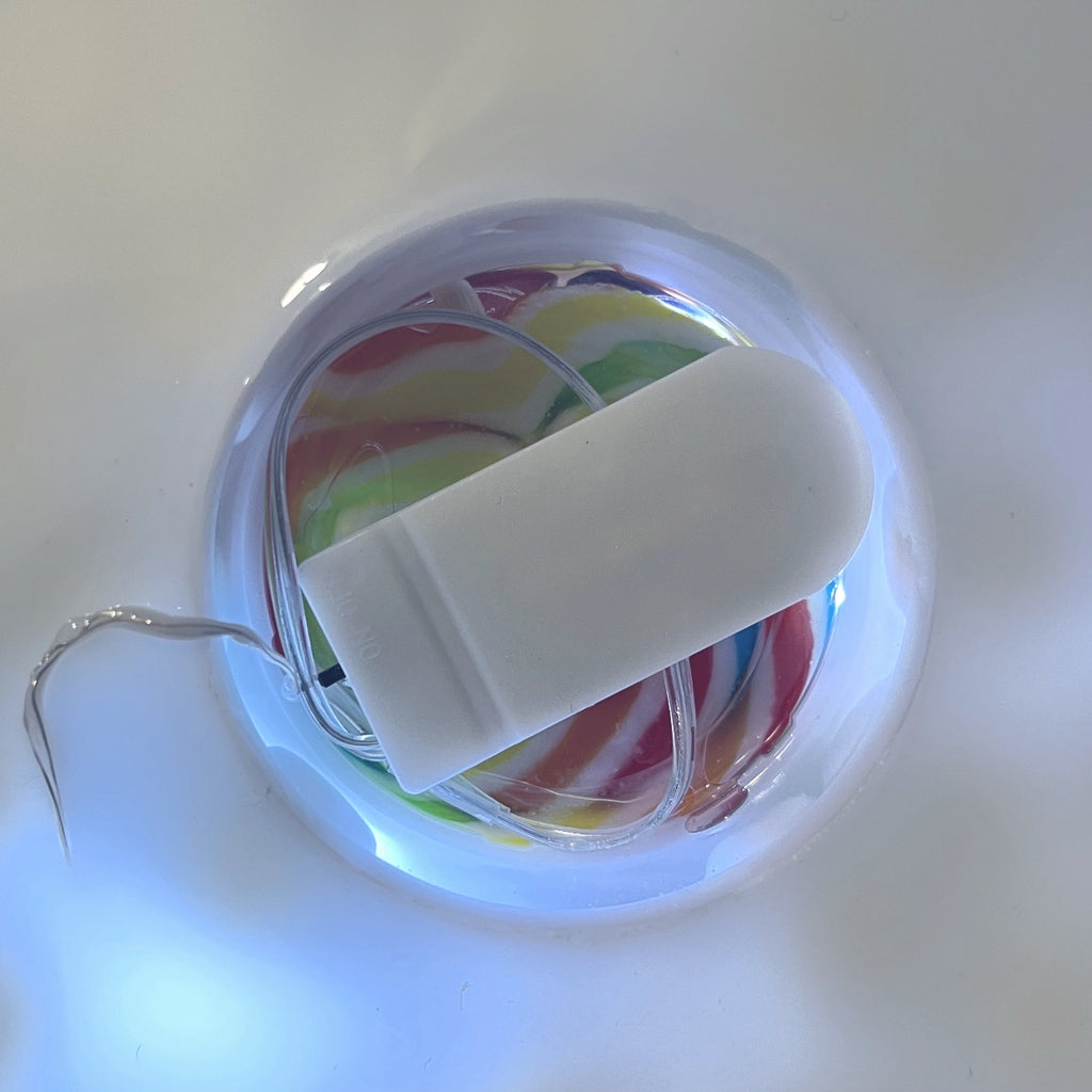 A close-up of a small white rectangular electronic component on a clear circular base, with thin metallic wires and a colorful marbled insert visible underneath.