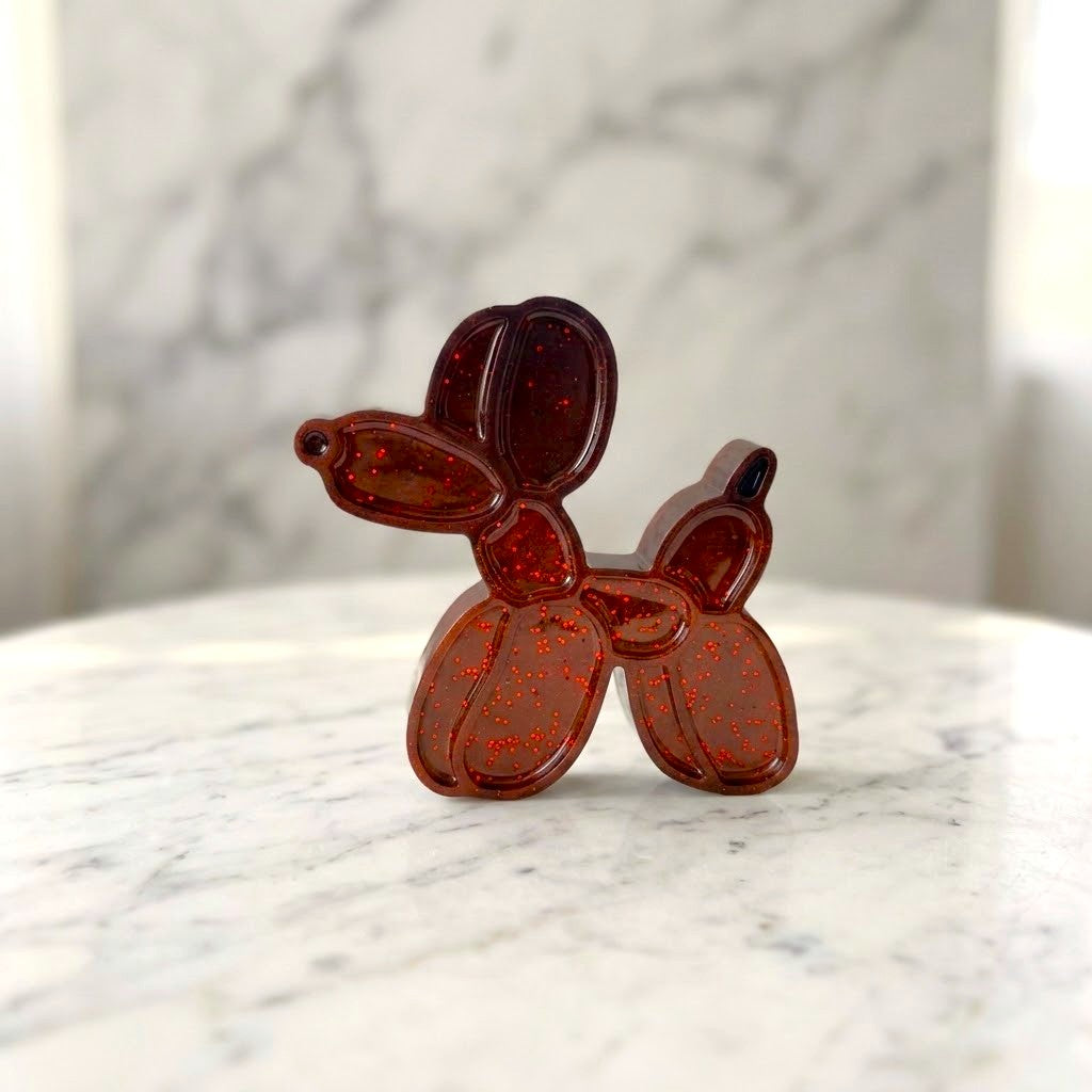 Balloon Dog Figurine - A small dog-shaped decorative object made of translucent reddish-brown material with red glitter, standing on a white marble surface against a softly blurred marble background.