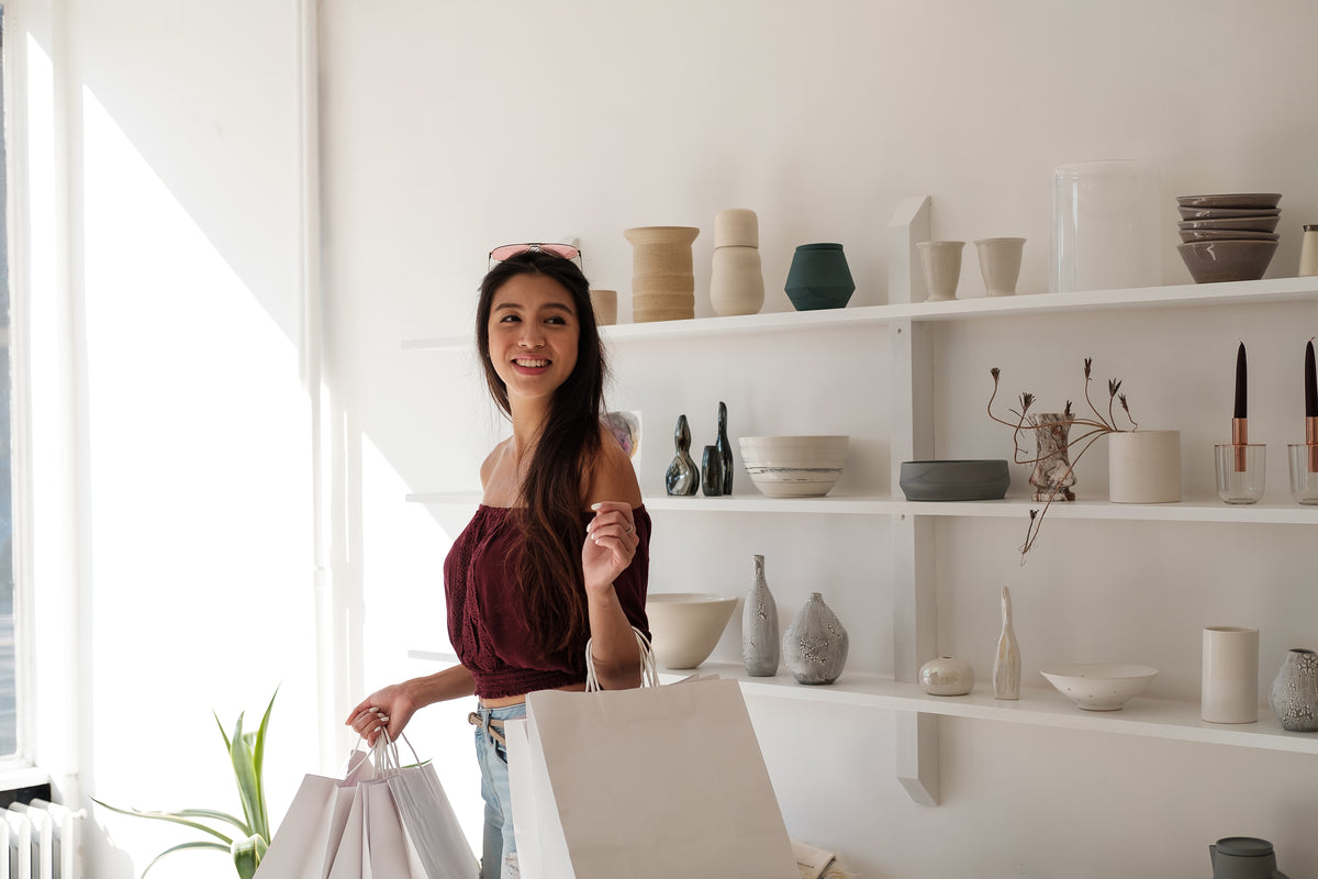 A woman smiling with shopping bags standing in front of three white shelves holding vases and ornaments 