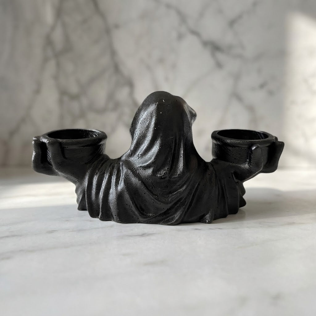 Grim Reaper Candle Holder - A black gothic-style candle holder shaped like a hooded figure holding two cups, displayed on a white marble surface with a matching marble background.