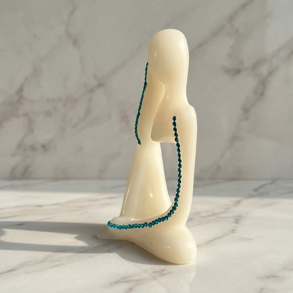 Nordic Resin Thinker - Side view of a white sculptural figurine of a seated female form with a beaded teal-green necklace draped around it, set on a white marble surface against a marble wall.