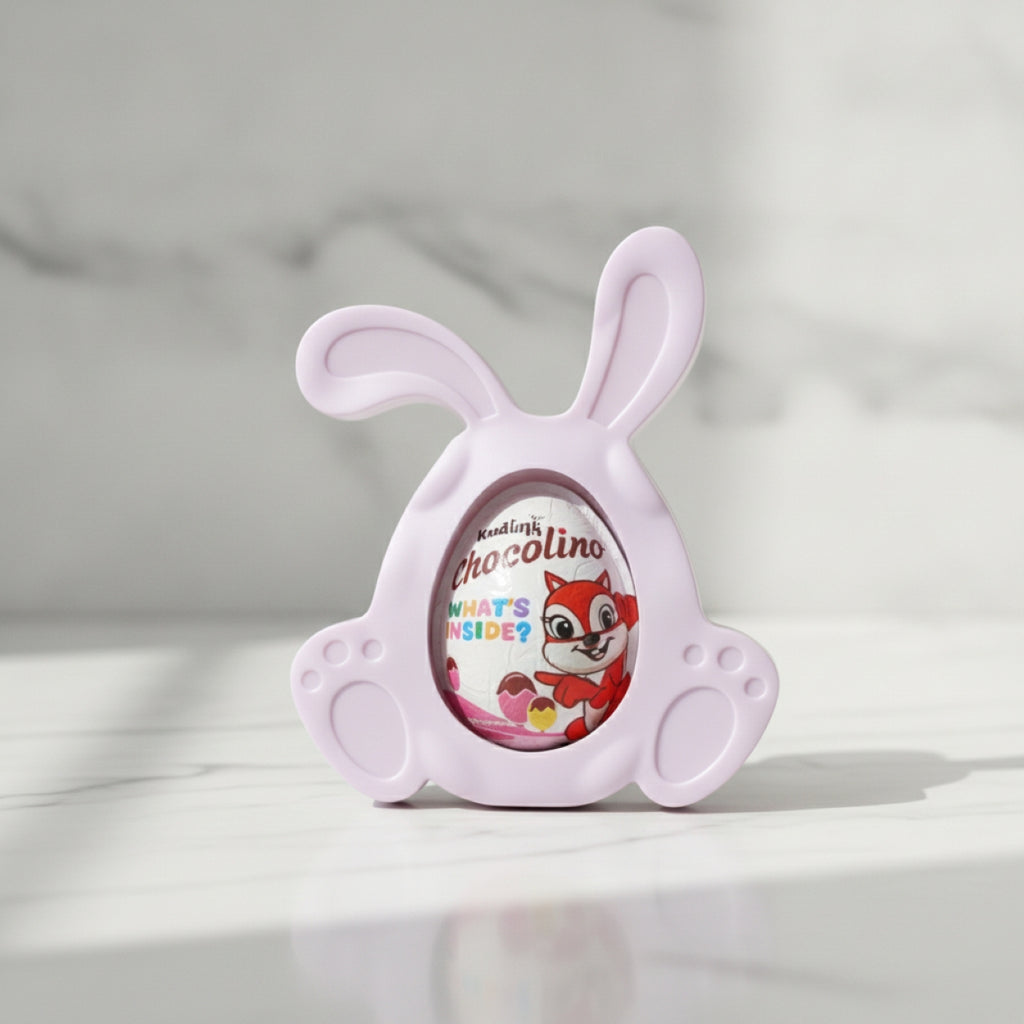A pastel pink bunny-shaped resin chocolate egg holder with a clear oval window showing a Chocolino chocolate egg inside, set on a light marble surface with a soft marble background.