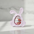 A pastel pink bunny-shaped resin chocolate egg holder with a clear oval window showing a Chocolino chocolate egg inside, set on a light marble surface with a soft marble background.
