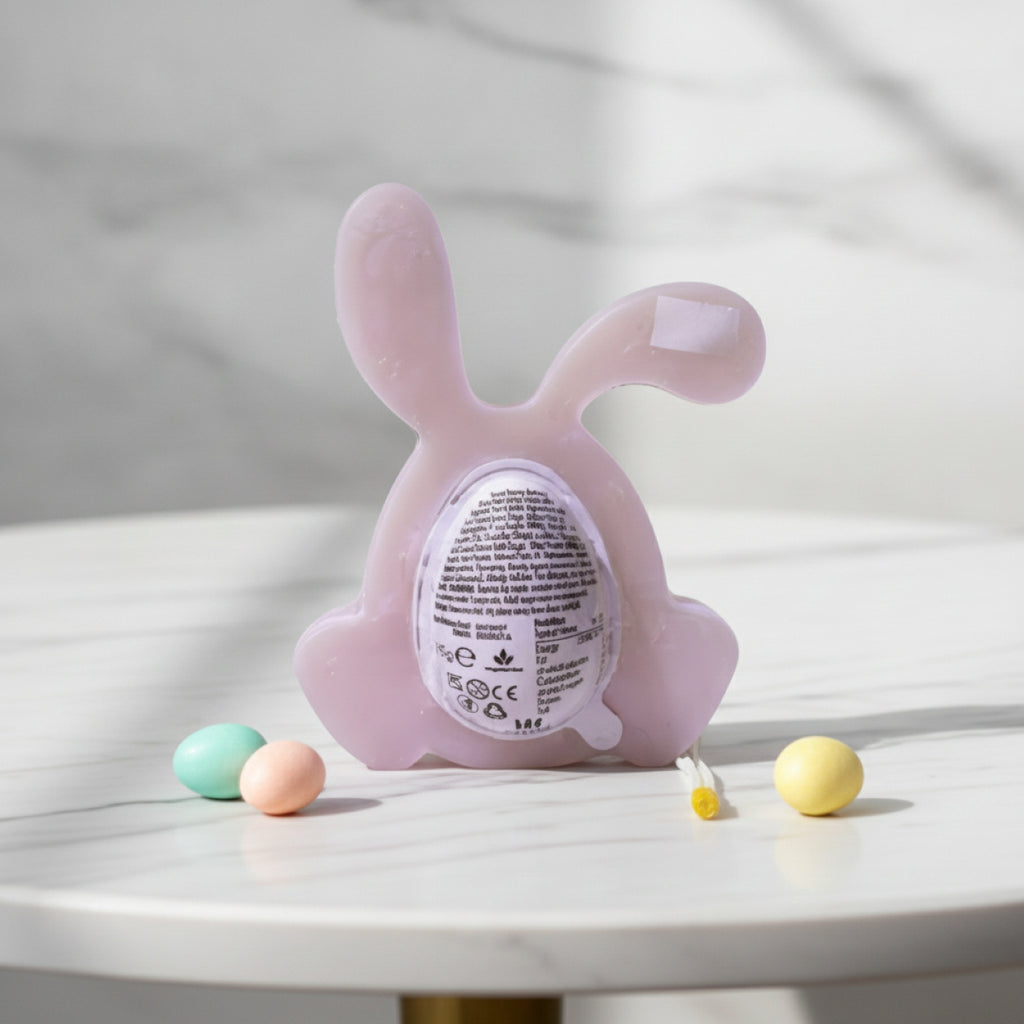 A pale pink bunny-shaped product with a circular egg and label on the back, displayed on a white marble surface with small pastel eggs around it.