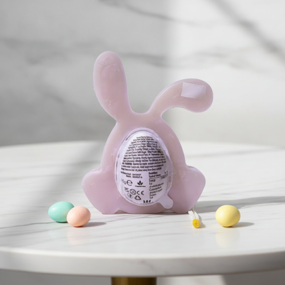 A pale pink bunny-shaped product with a circular egg and label on the back, displayed on a white marble surface with small pastel eggs around it.
