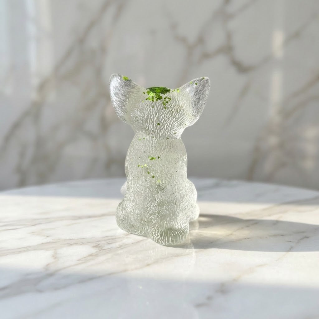 Chihuahua Dog Figurines - Back view of a small clear resin figurine shaped like a seated dog, with tiny green inclusions near the top, photographed on a white marble surface against a softly blurred marble background.