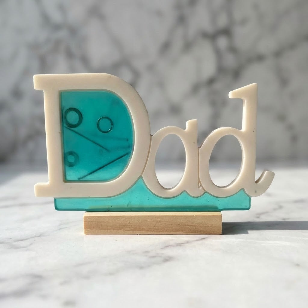 Handmade Dad Plaque - A decorative 3D letter sign spelling 'Dad' in cream, mounted on a turquoise translucent panel and a small wooden base, photographed on a marble surface with a marble background.