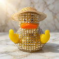 Diamante Duck Figurine - A rhinestone-covered rubber duck figurine wearing a straw hat, with yellow fingers pointing upwards and an orange beak, displayed on a light marble surface.
