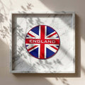 Union Jack Coasters - A circular England-themed Union Jack coaster with a distressed red, white, and blue design and the word 'ENGLAND' across the centre, displayed inside a white marble shadow box frame on a beige wall with leaf shadows.