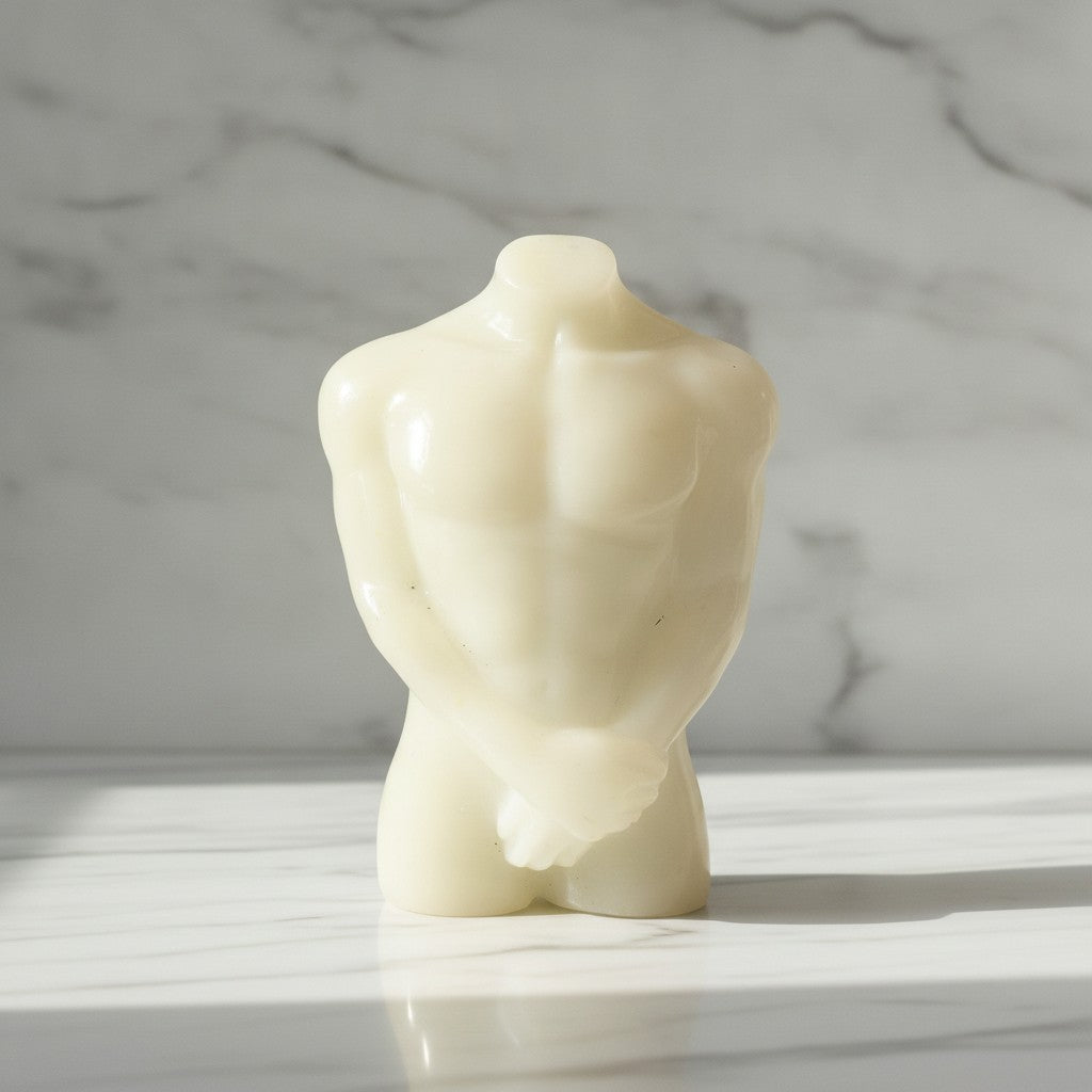A small sculptural male torso figurine in a warm white resin material, standing on a white marble surface with a gray marble background
