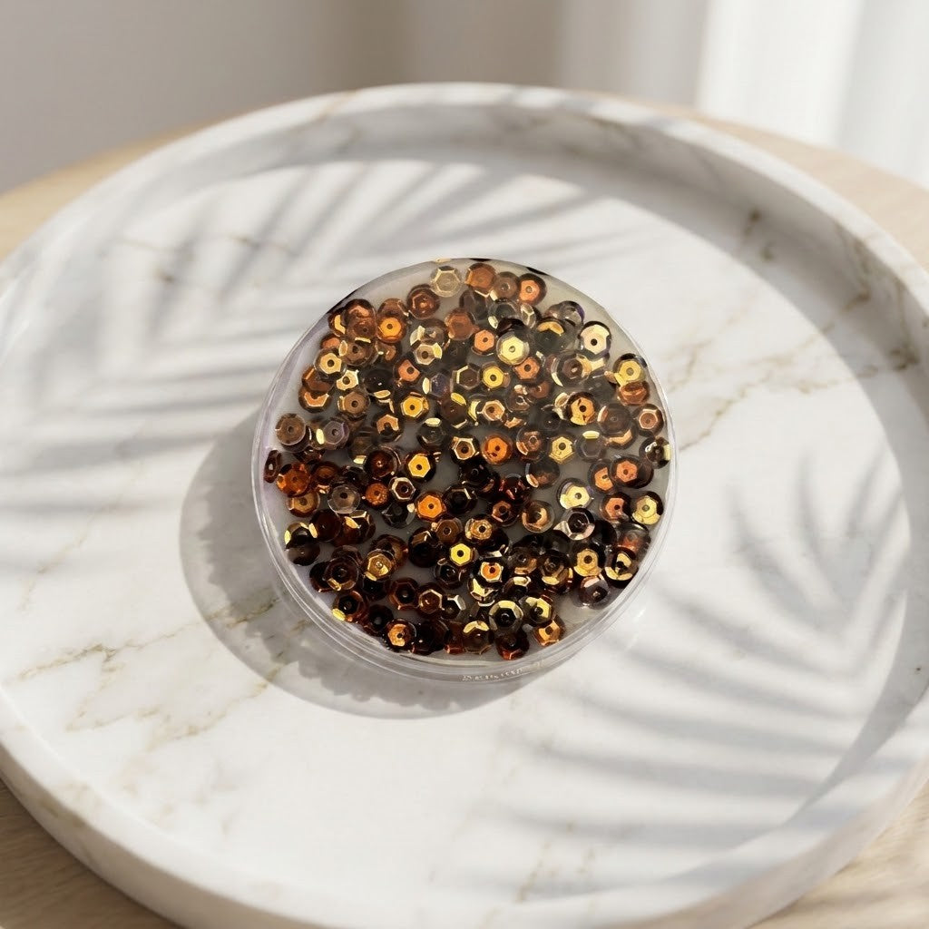 Golden Sequin Coasters - A round coaster filled with small metallic sequins in gold, bronze, and dark brown tones, placed on a white marble tray with leaf shadows cast across it.