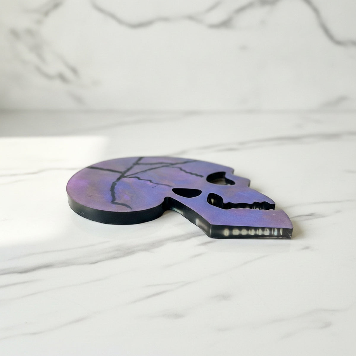 Skull shaped coasters with a blue, purple, and pink marbled pattern and black crackle lines, displayed on a white marble tray with leaf shadows cast across it.