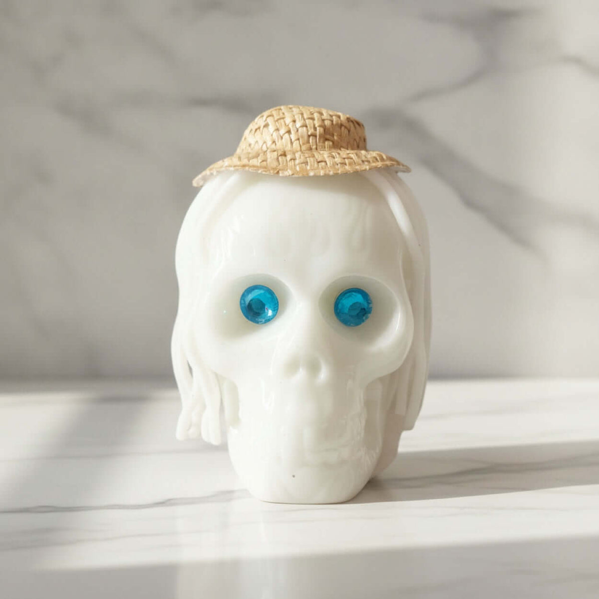 A white skull figurine with textured hair and a small woven straw hat, featuring bright blue gem-like eyes, set on a light marble surface against a marble background.