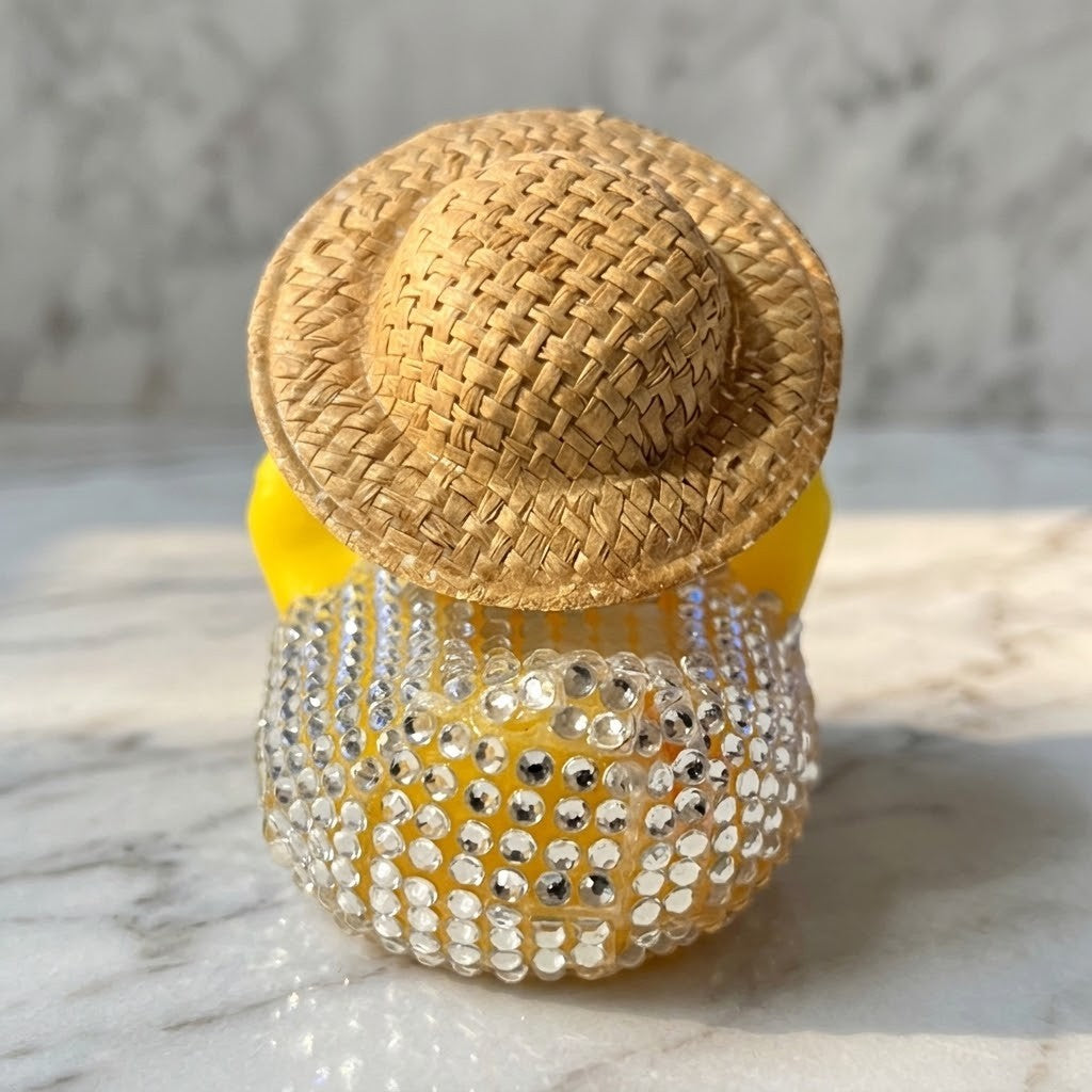 Diamante Duck Figurine - Back view of a  small decorative figurine shaped like a yellow duck character wearing a woven straw hat, photographed on a light marble surface with a marble background.