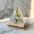 Christmas Tree Pyramid Lamp - A clear resin pyramid snow globe featuring a small penguin wearing a blue hat, a miniature Christmas tree, and gold glitter accents on a white base, set on a marble surface.