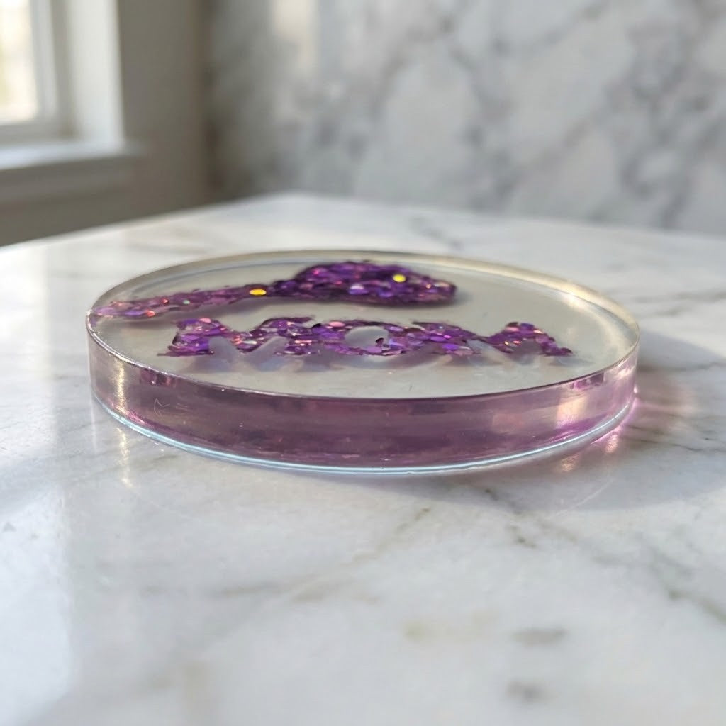 Handmade MOM Coaster - A round clear resin coaster with a translucent purple base and purple glittery lettering, placed on a white marble surface.