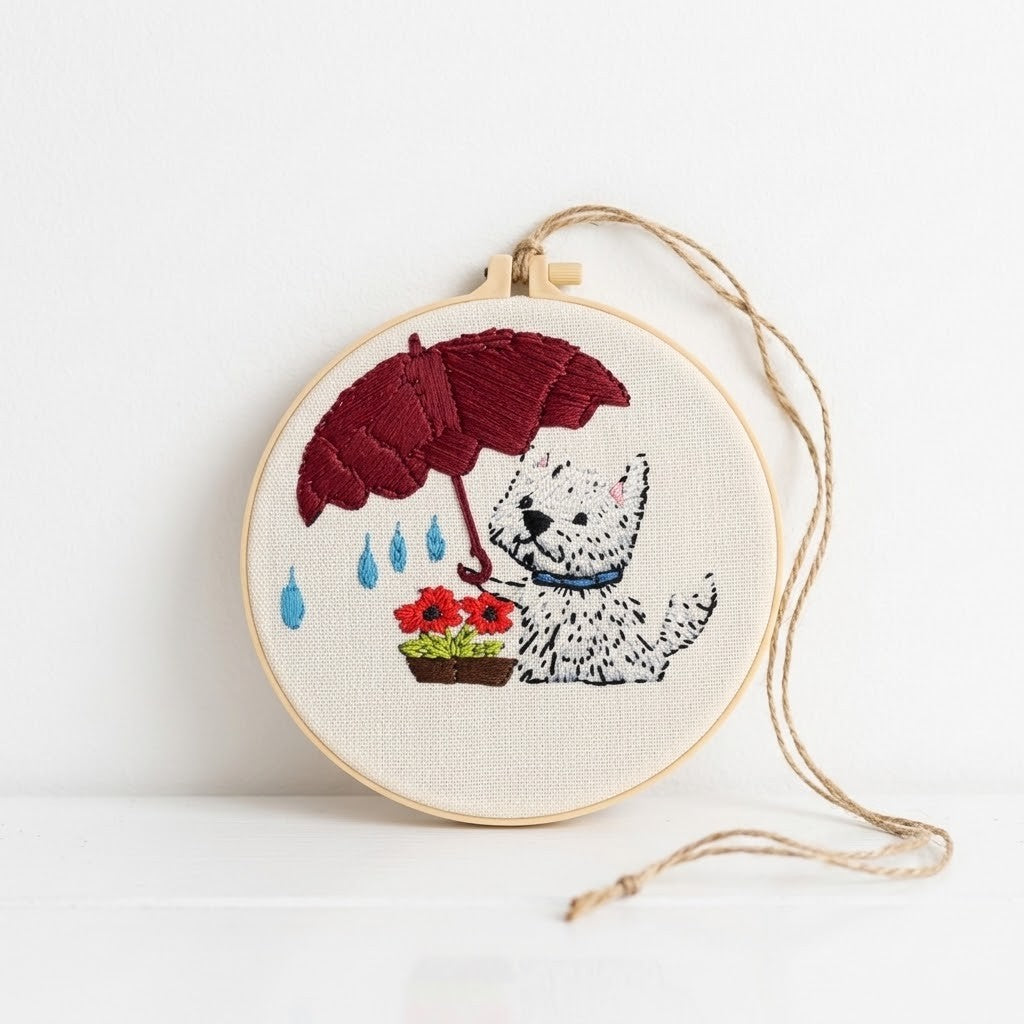 Dog Wall Art - A circular embroidery hoop with a finished cross-stitch design of a white dog under a dark red umbrella, holding red flowers, with a hanging twine loop, set against a plain white background.