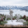 Reindeer Gift Set - Two silver rhinestone reindeer figurines, one standing and one lying down, decorated with bows and set on a snowy wooden surface with pine branches, pinecones, and a blurred mountain and frozen lake background.