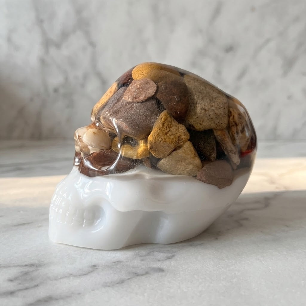 Gothic Resin Stone Skull - Side view of a polished white skull-shaped object filled with smooth river stones in earthy brown, tan, and grey tones, photographed on a light marble surface with a soft marble background.