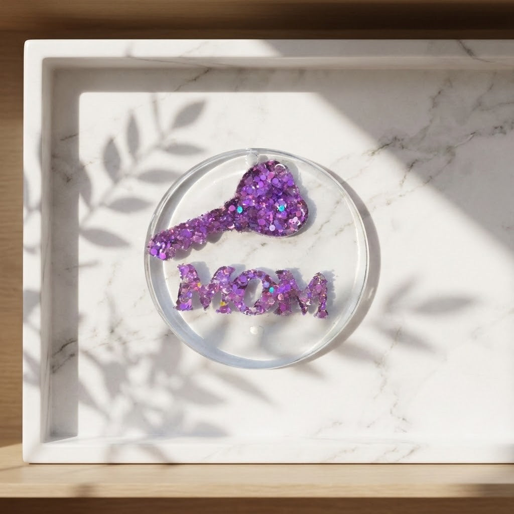 Handmade MOM Coaster - A white marble tray holding a purple glitter coaster  and matching glitter letters spelling 'MOM', set on a white marble surface with leaf shadows from nearby foliage.