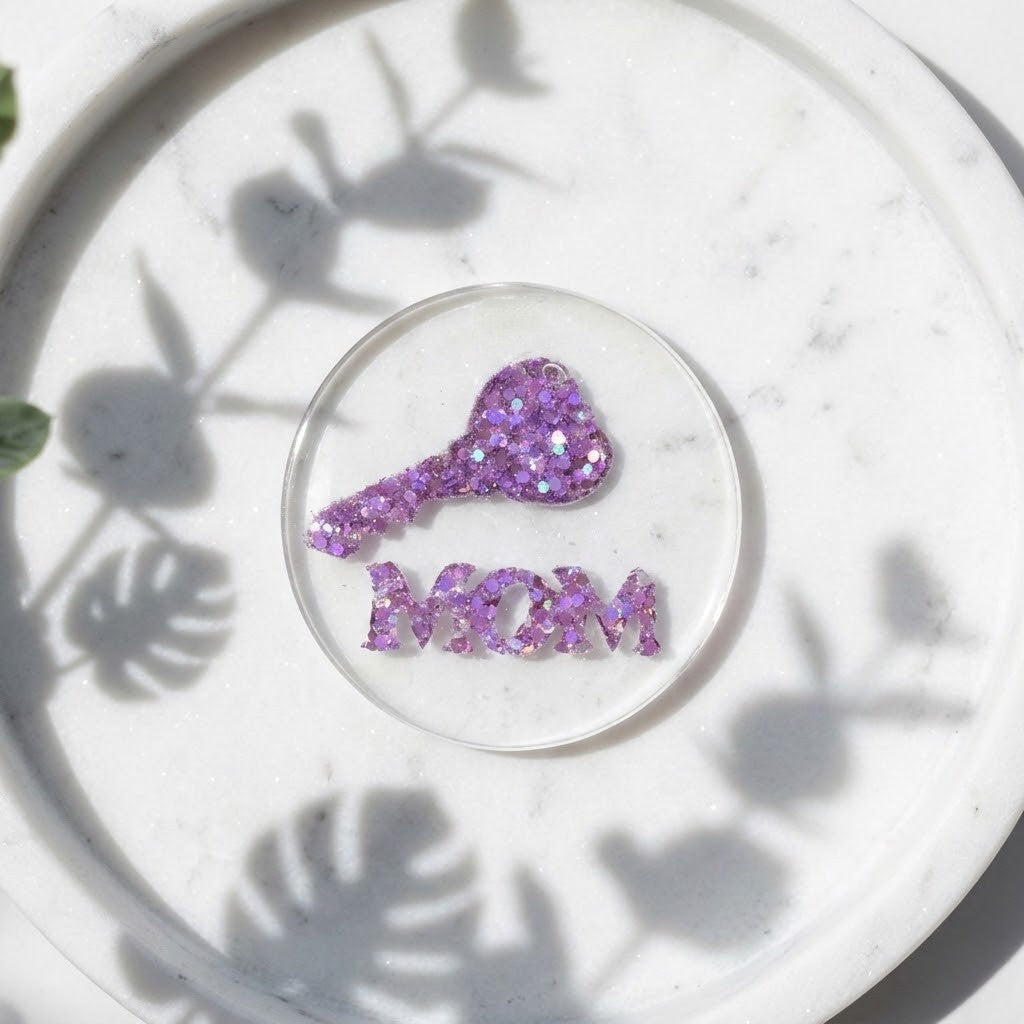 Handmade MOM Coaster - A clear circular resin coaster featuring a purple glitter key and the word 'MOM' in matching purple glitter letters, displayed on a white marble tray with leaf shadows.