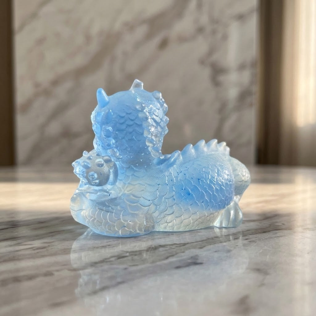 Dragon Figurines - Back of a small transparent blue dragon figurine with dark blue eyes sits on a white marble surface, against a matching marble wall, holding a blue baby dragon and egg in its front paw