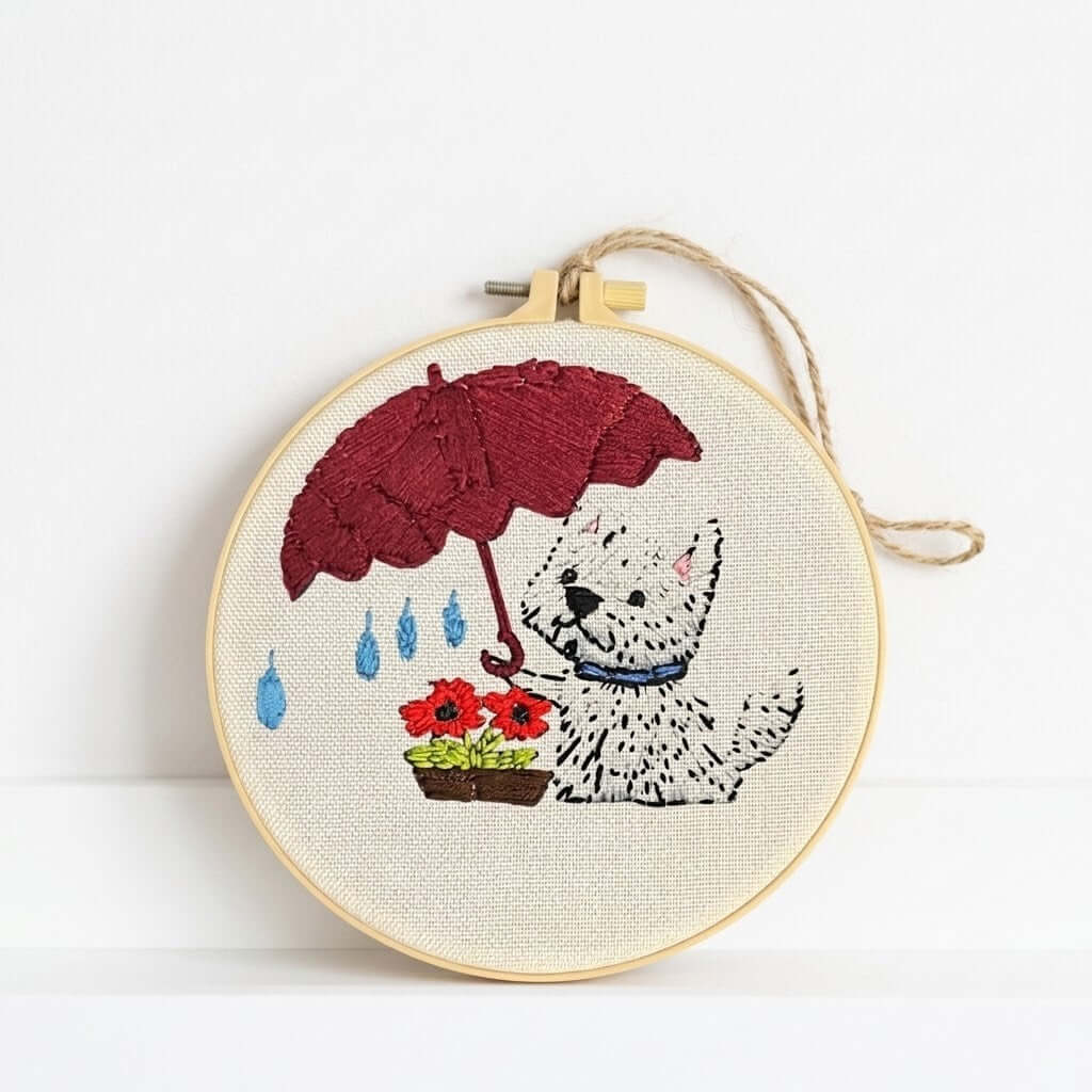 Dog Wall Art - A circular embroidery hoop with a finished cross-stitch design of a white dog under a red umbrella next to flowers, hanging on a white wall.