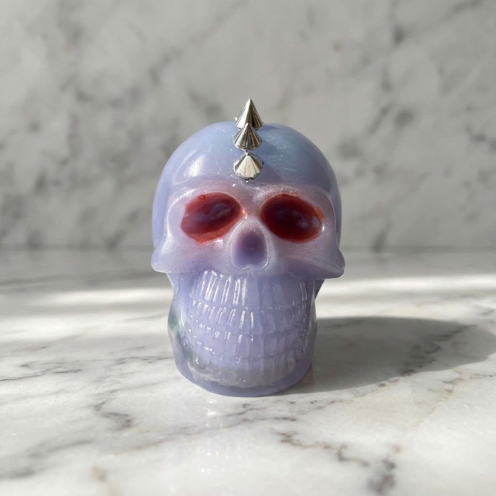 Studded Gothic Skull - A small skull-shaped figurine, lavender purple with dark red eye sockets and a silver spikes on top, photographed on a white marble surface against a marble background.
