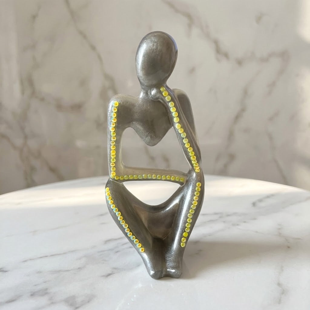 Nordic Resin Thinker Figurine - A modern abstract figurative sculpture of a seated human form in a metallic silver finish, accented with small yellow beads along the spine, arms, and legs, displayed on a white marble surface against a marble wall background.